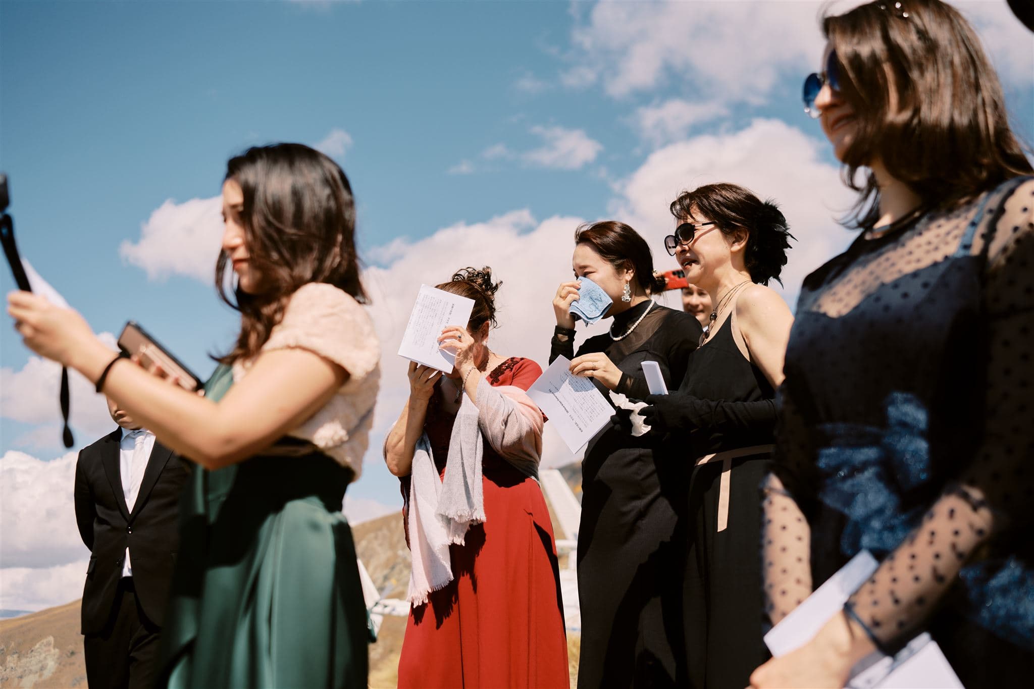 Queenstown New Zealand Heli Wedding Elopement Photographer クイーンズタウン　ニュージーランド　エロープメント 結婚式 | A group of formally dressed women gathered outdoors, one wiping tears with a tissue, as the Wedding Photographer captures every moment.